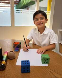 Niño sonríe con un lápiz en la mano junto a dos dados grandes sobre la mesa de trabajo.