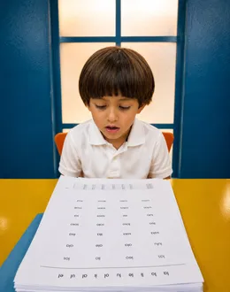 Niño lee sílabas en voz alta frente a una hoja de lectura en el aula.