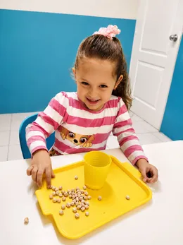 Niña sonriente clasifica garbanzos sobre una bandeja amarilla en un ejercicio de motricidad fina.