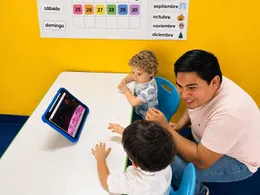 Terapeuta trabaja con dos niños pequeños frente a una tablet, bajo el calendario del aula.