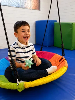 Niño riendo sentado en el columpio de plataforma de la sala de integración sensorial.